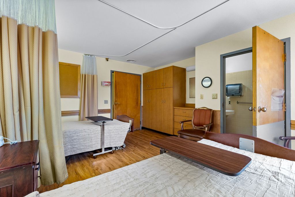 Quality Life Services room with bed, privacy curtain, wooden furniture, and doorway to bathroom.