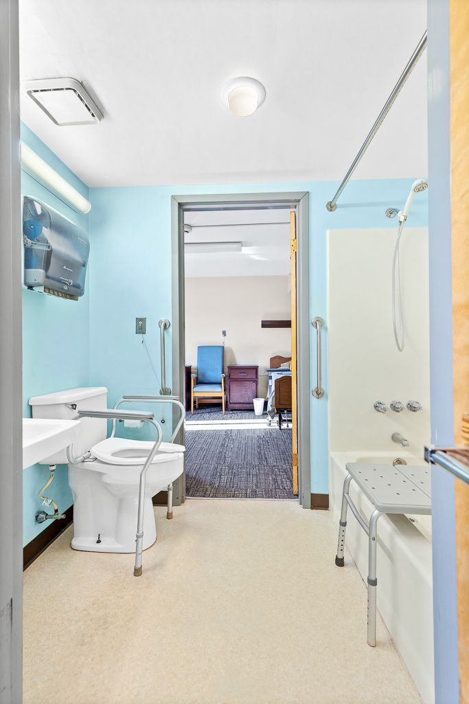 Accessible bathroom with toilet, sink, and bathtub. Light blue walls, grab bars, open doorway to another room.