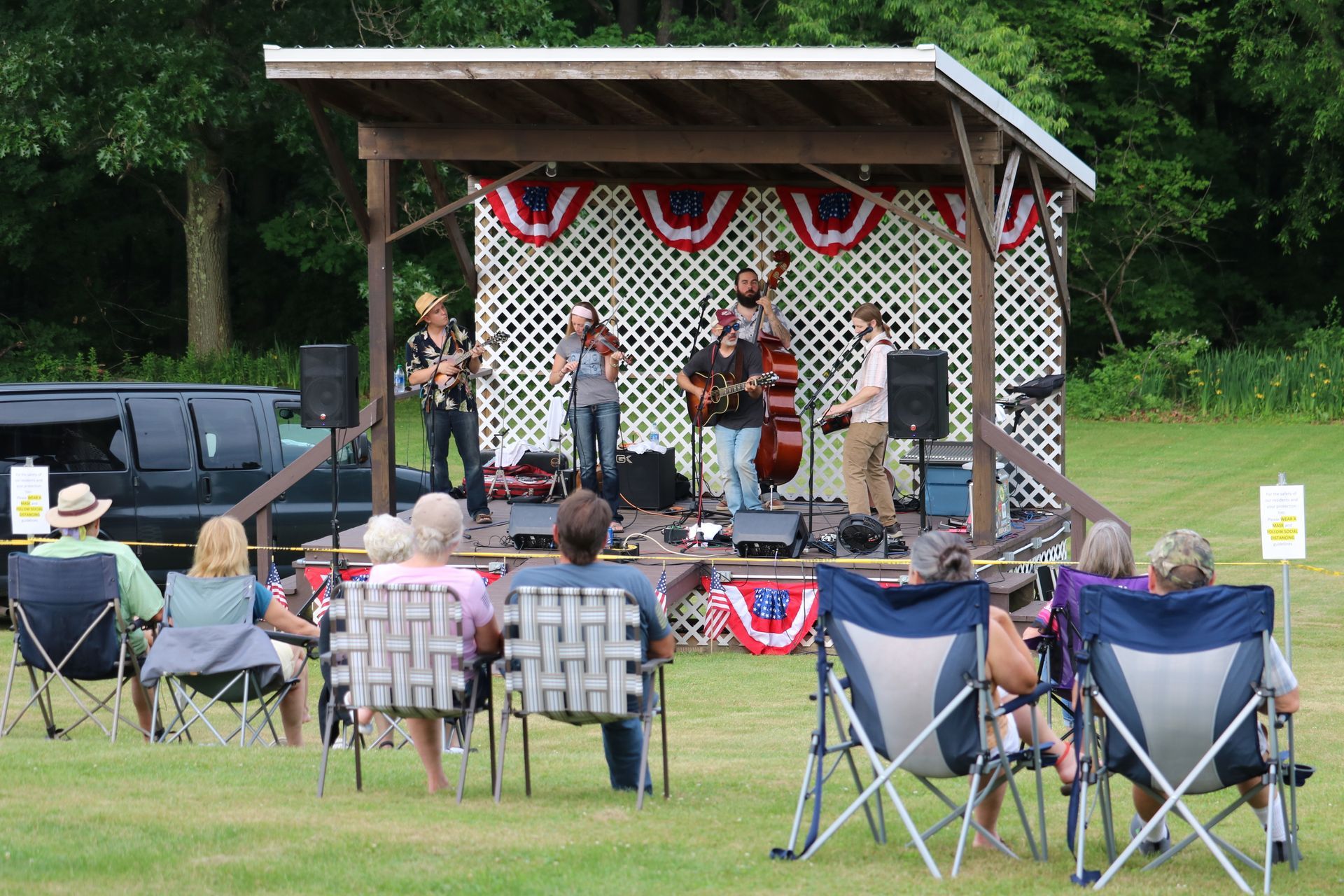 Bluegrass band performing on outdoor stage, audience seated in folding chairs.