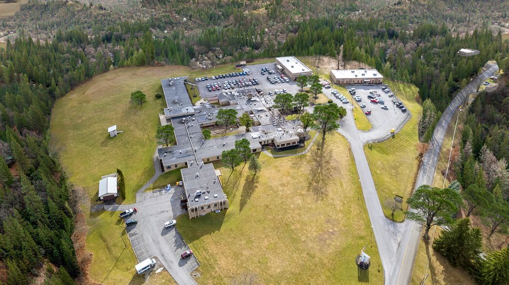 Aerial view of a long, low building complex with parking lots, on a grassy hill surrounded by trees.