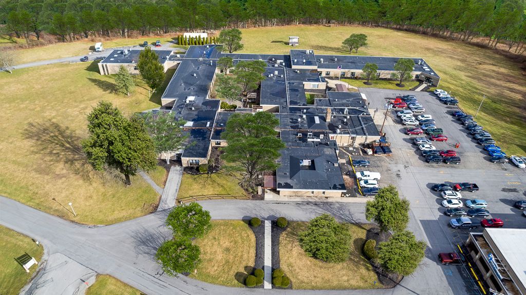Aerial view of a long, low, rectangular building with a dark roof and a parking lot. Trees and grass surround it.