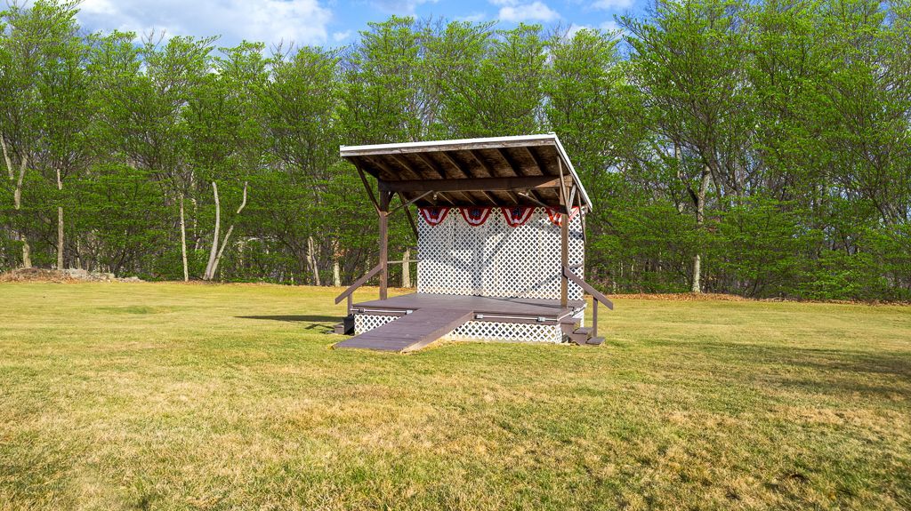 A small, wooden stage with a sloped roof sits on a grassy field, trees in the background, blue sky.