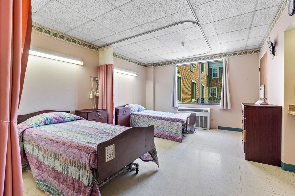 Two hospital beds in a room with a window, curtains, and dresser. Soft pink walls.