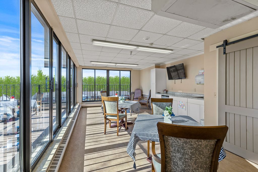 Sunlit room with tables, chairs, and large windows overlooking a green landscape. 
