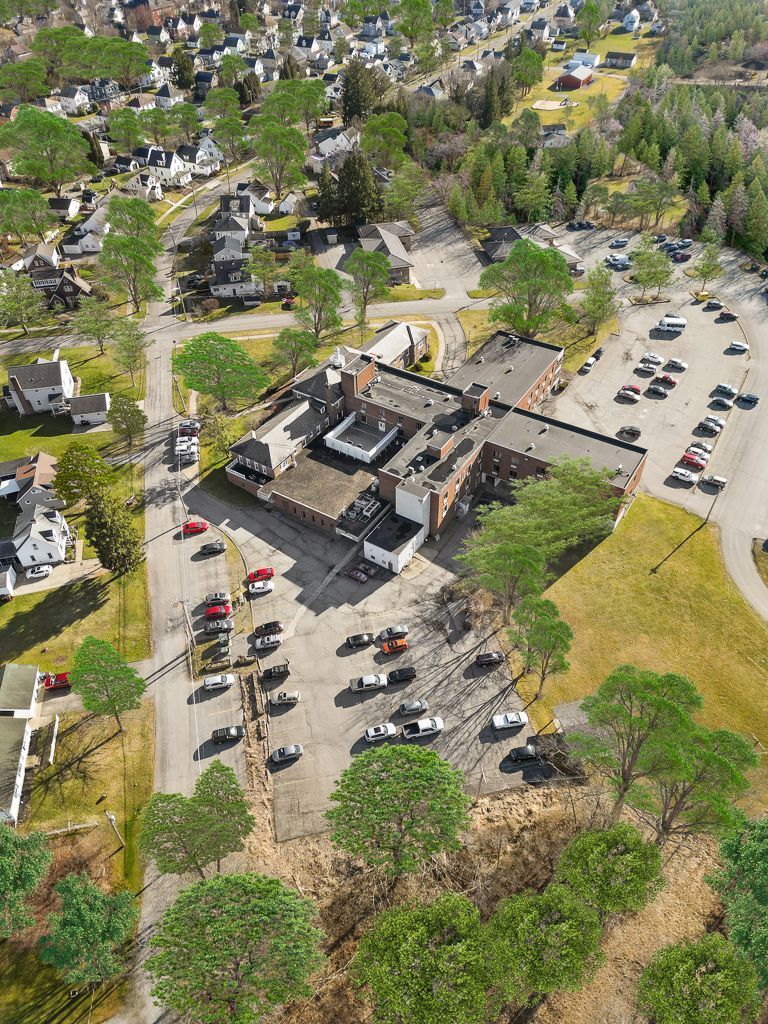 Aerial view of a building surrounded by cars in a parking lot and trees; residential neighborhood in the background.
