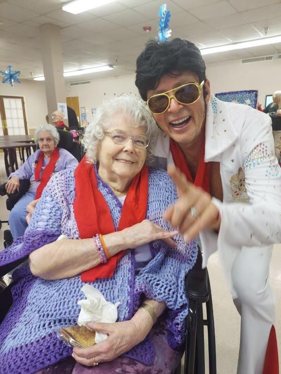 Elvis impersonator smiles with a woman in a wheelchair, indoors. They both wear red scarves, she holds a napkin.