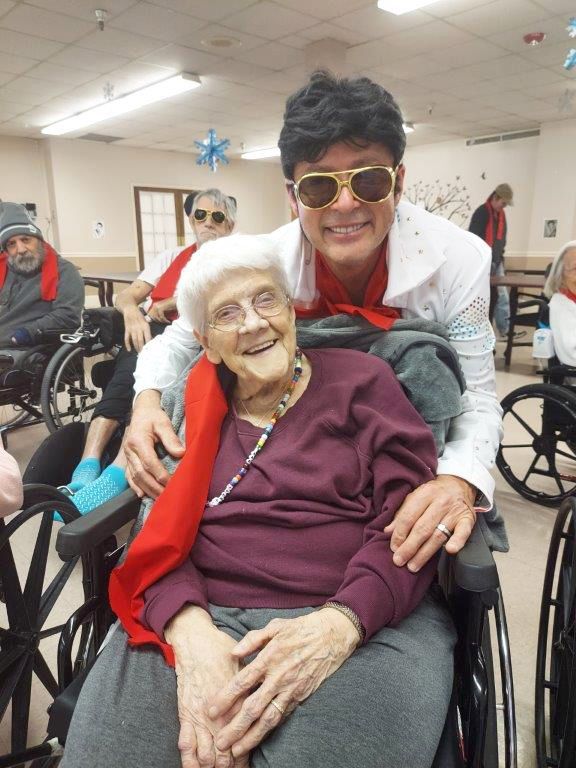 Elvis impersonator with elderly woman in wheelchair, smiling, in a community room.