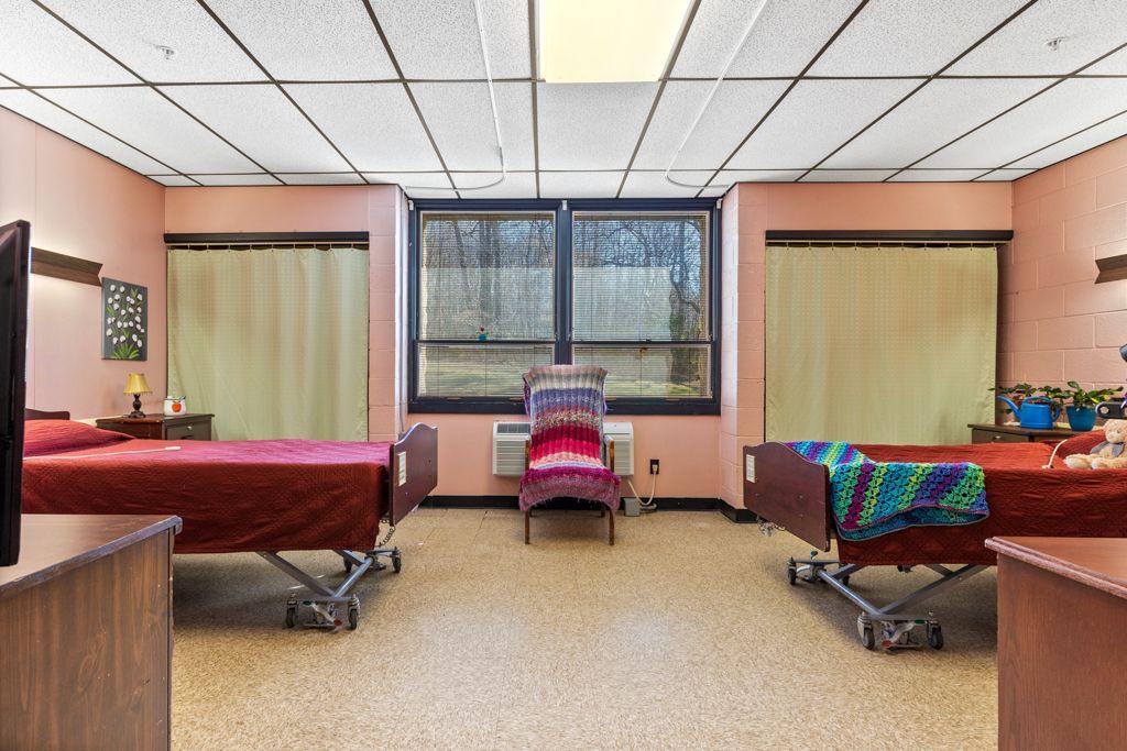 Two hospital beds in a room with a window, beige floor, pink walls, and a chair.