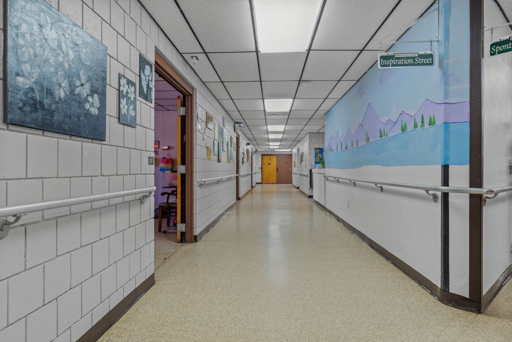 Long, empty school hallway with white tiled walls, handrails, and artwork. A door is open, showing a glimpse of a room.