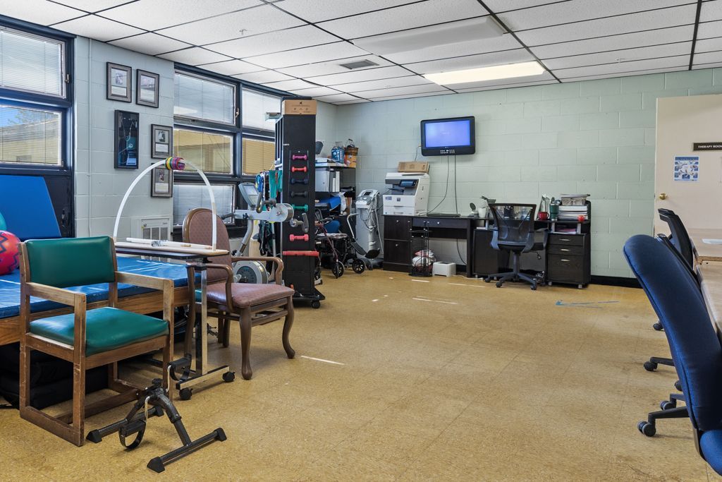 Interior of a room with furniture, medical equipment, and office supplies. Windows on left, door on right.