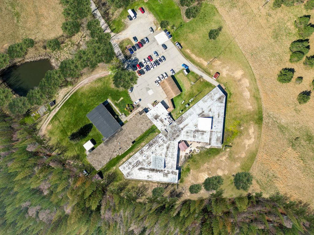 Overhead shot of a building with a parking lot and small pond surrounded by fields and trees.