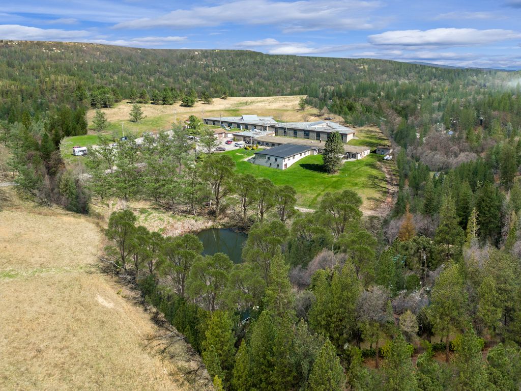 Aerial view of a long, low building in a clearing surrounded by trees, with a pond in the foreground.