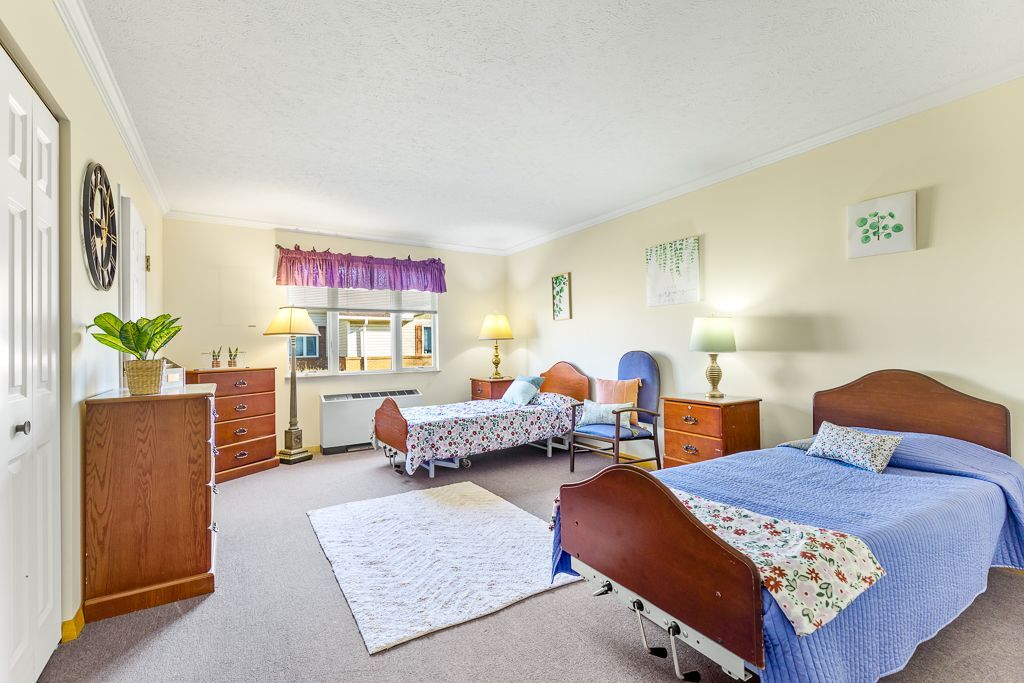 Spacious, well-lit senior living room with two beds, two chairs, and various furnishings. Beige walls, carpeted floor.