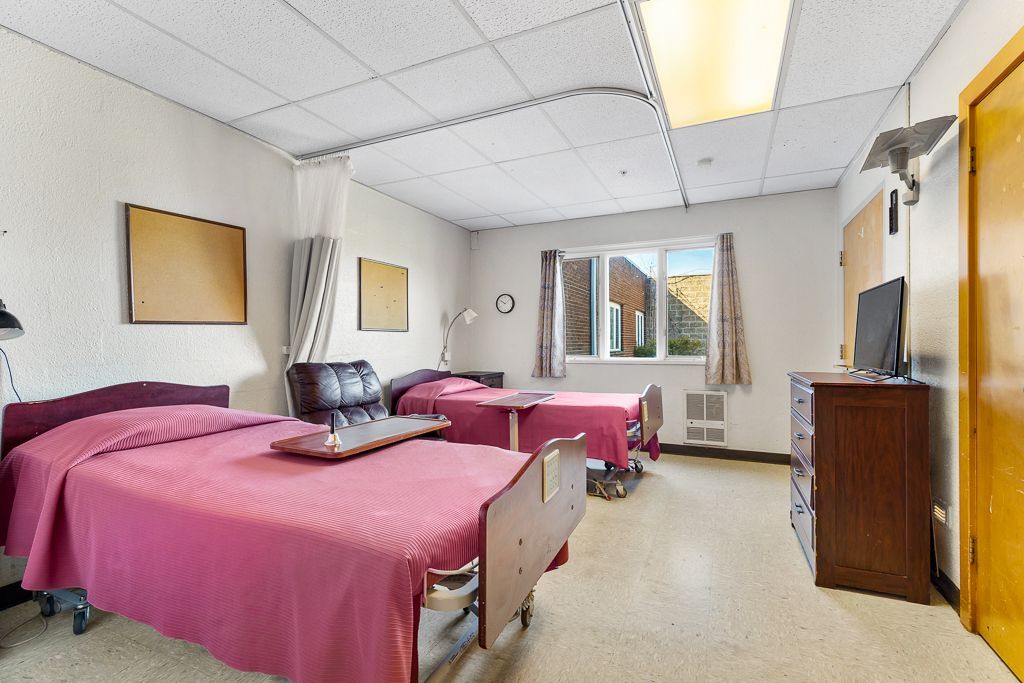Two hospital beds with pink linens in a shared room, small window, wood door, and dresser.