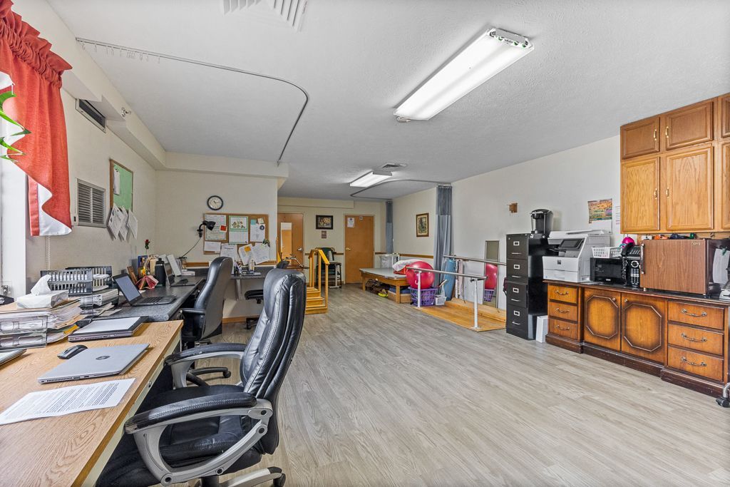 Office interior with desks, cabinets, and various equipment. Brightly lit with a window and chairs.