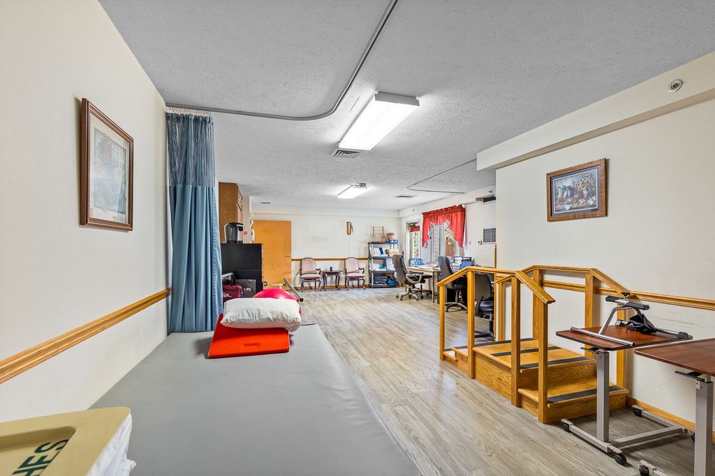 Rehab room with wooden flooring, exercise equipment, and seating. Beige walls, fluorescent lights.