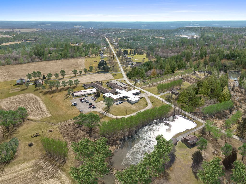 Aerial view of a complex with buildings, parking, fields, and a road surrounded by trees and water.