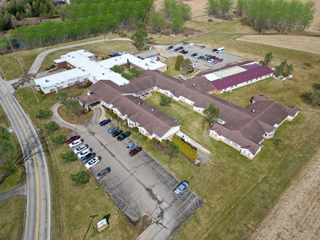 Aerial view of a long, white building with a cross-shaped section, parking lots, and a road, surrounded by greenery.