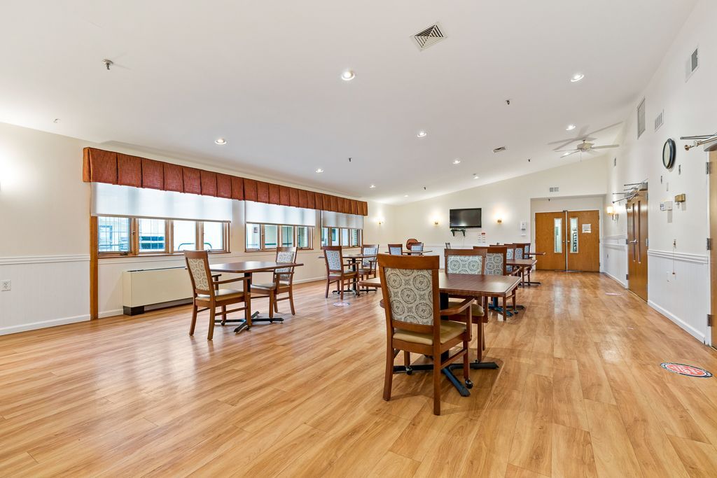 Empty dining area with tables, chairs, and wood flooring. Features windows, doors, and overhead lighting.