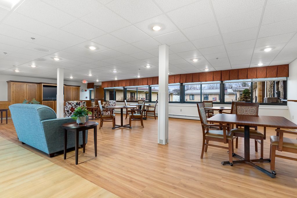 Spacious common room with tables, chairs, sofa, and large windows. Warm wood floors and neutral colors.