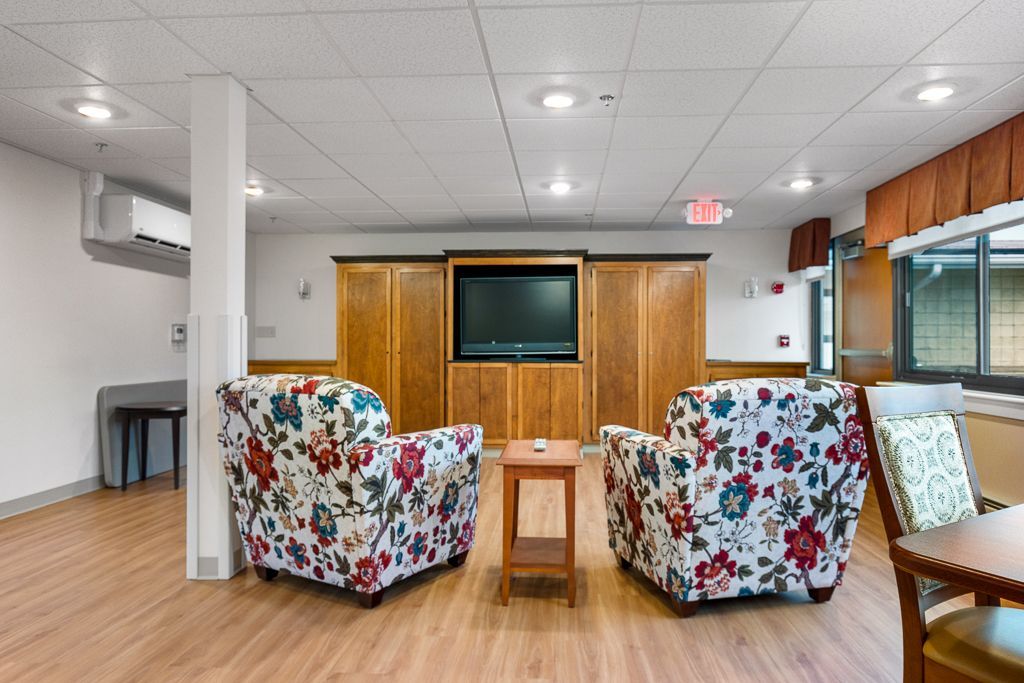 Two floral armchairs face a TV in a sitting room with wood cabinets, wood floor, and large windows.