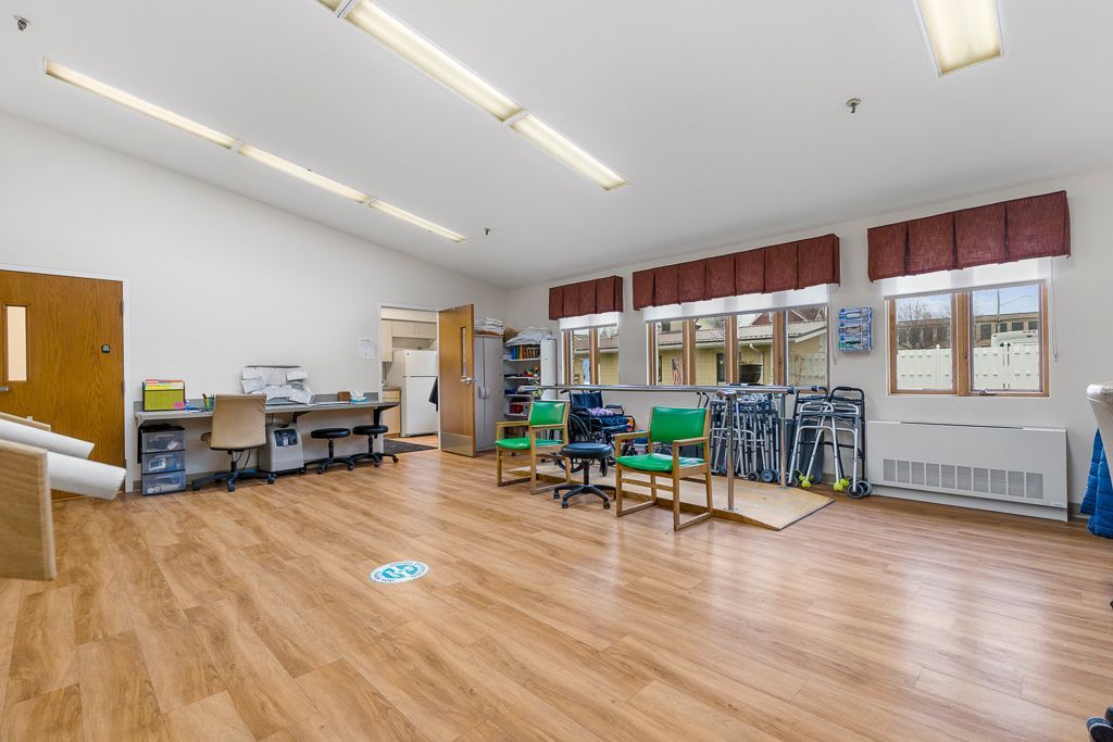 Large, physical therapy room with wood-look floor, fluorescent lights, and windows with brown valances.