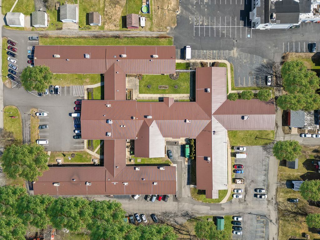 Overhead view of a brown-roofed building with a cruciform layout surrounded by parking, trees, and small buildings.