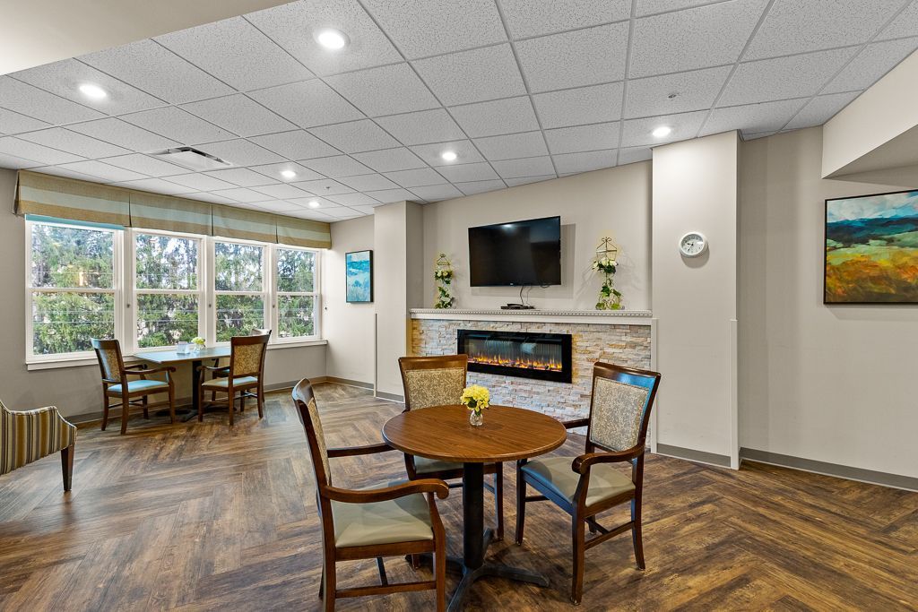 Cozy lounge with fireplace, TV, round tables, chairs, and large windows. Brown and beige tones, bright ceiling lights.