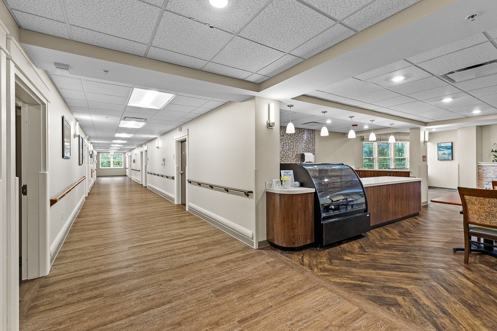 Hallway in a care facility with doors, a cafe counter, and seating. Beige walls and patterned flooring.