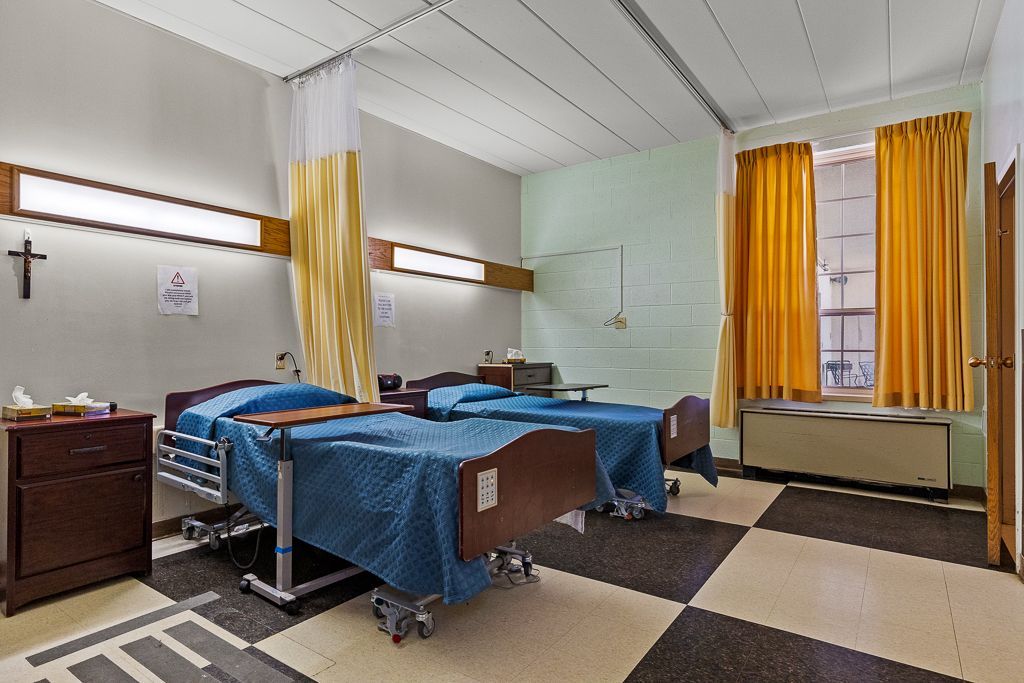 Hospital room with two beds, blue covers, and yellow curtains. Checkerboard floor.