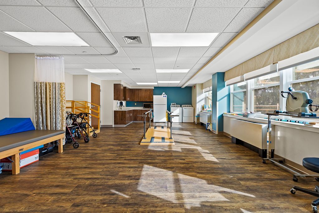 Rehab room with exercise equipment, treatment tables, and windows; brown flooring, beige and blue walls.