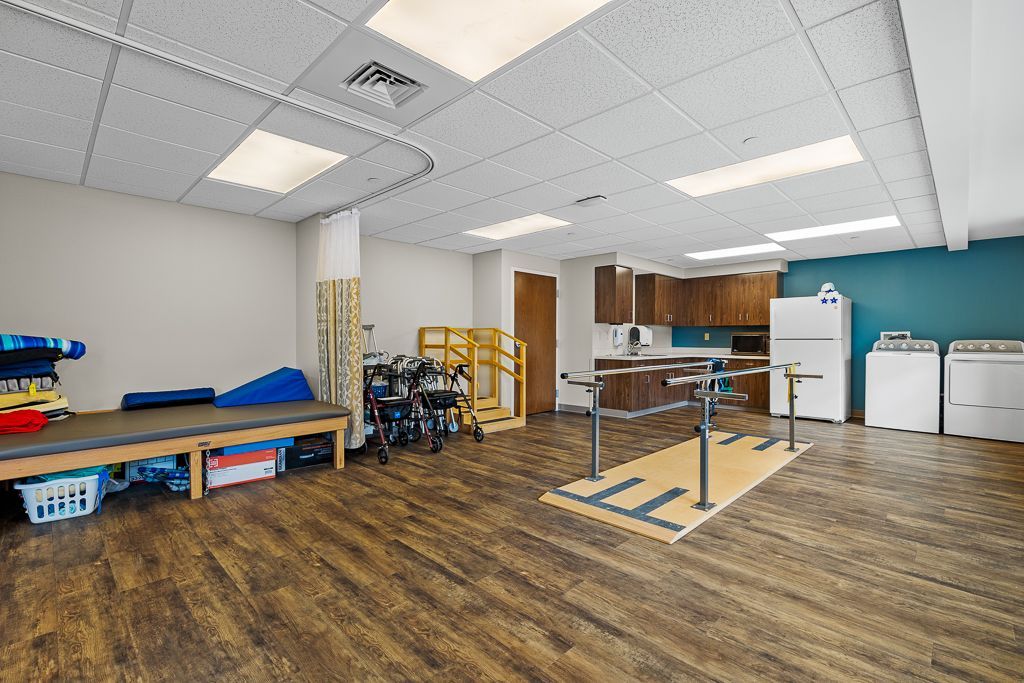 Rehabilitation room with various equipment, including parallel bars, a mat table, and a walker. Brown wood flooring.