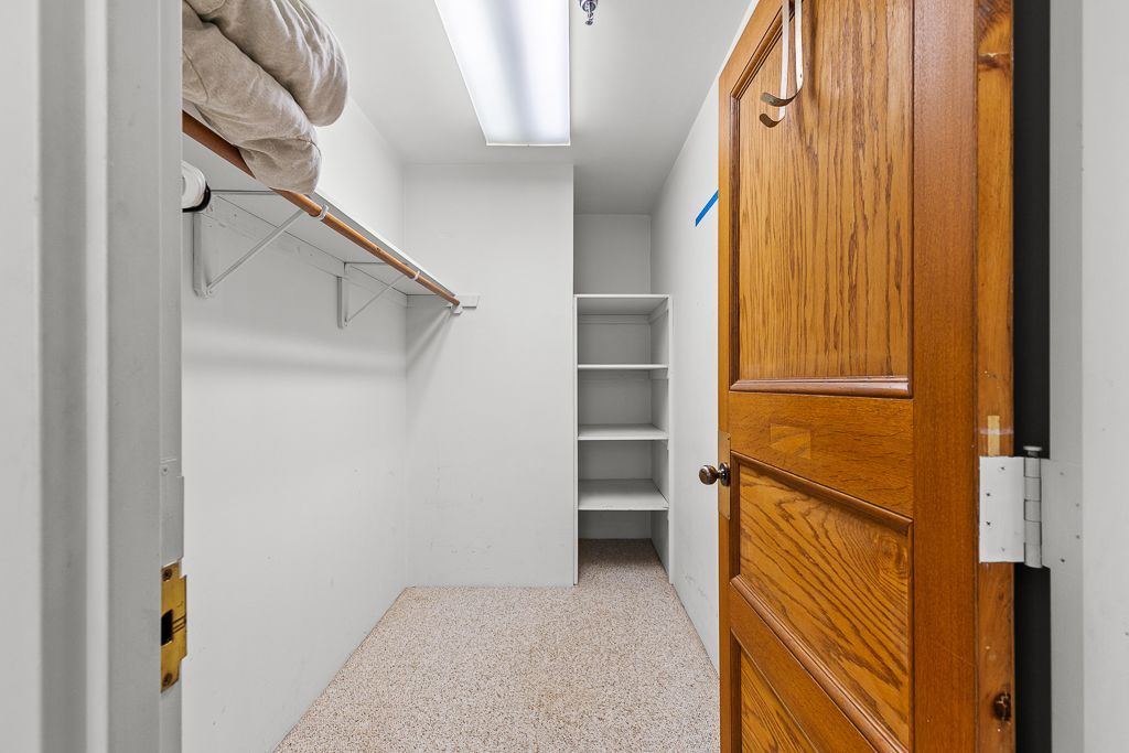 Walk-in closet with open wooden door, shelving, and tan speckled flooring.