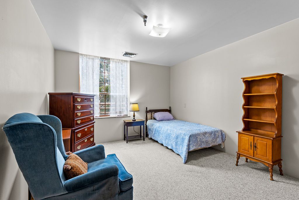 Bedroom with blue armchair, single bed, two wood dressers, and a window with white curtains.