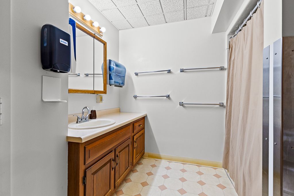 Bathroom with wood vanity, mirror, towel racks, and shower curtain. Tile floor has a tan pattern.