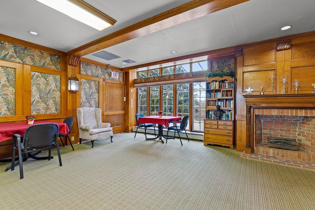 A wood-paneled room with tables, chairs, a fireplace, and large windows overlooking trees.