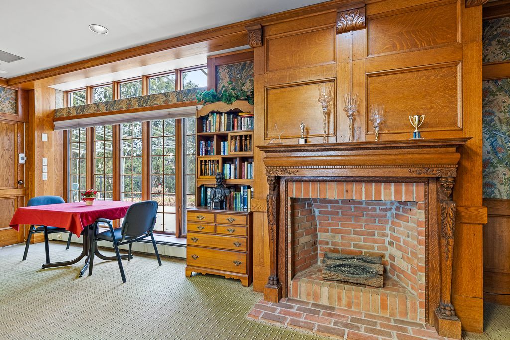 Cozy room with fireplace, built-in bookshelf, table with red tablecloth, and large windows looking out on trees.