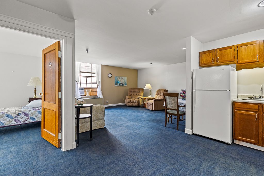 Open-plan living space with kitchen, chairs, and bedroom visible through an open door.