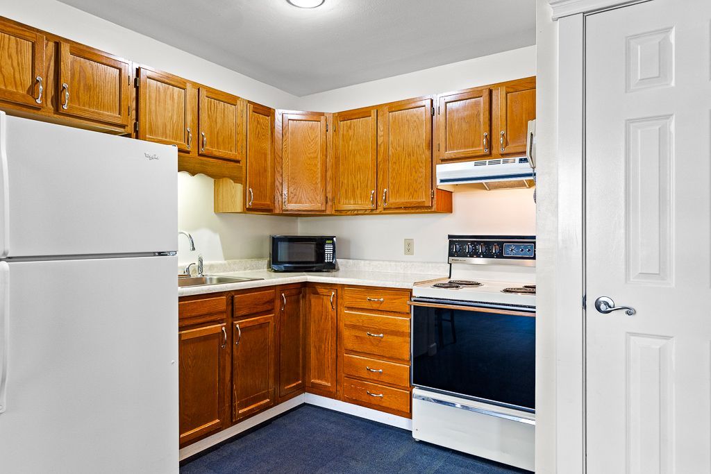 A small kitchen with light-colored wooden cabinets, white appliances, and a closed white door.