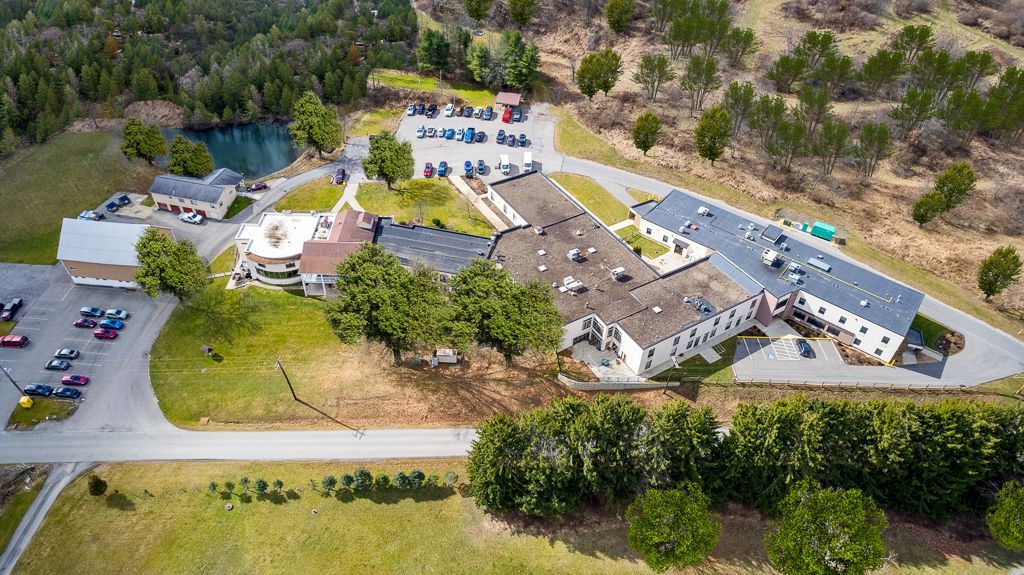 Aerial view of a complex with buildings, parking, and a small pond, surrounded by trees and a field.