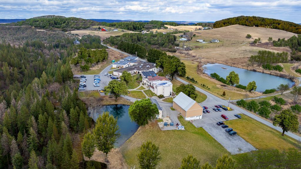 Aerial view of a complex with buildings, parking, and ponds surrounded by forest and fields.