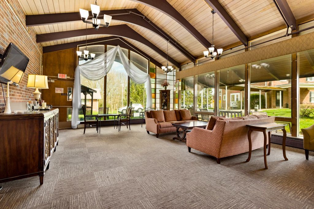 Lounge with large windows, sofas, and tables. Brown ceiling beams, brick wall, and carpet flooring.