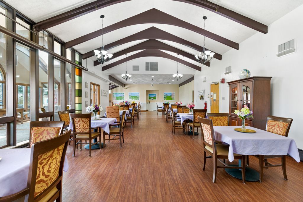 Dining room with tables, chairs, and large windows. Wooden floor, brown beams, and light blue tablecloths.