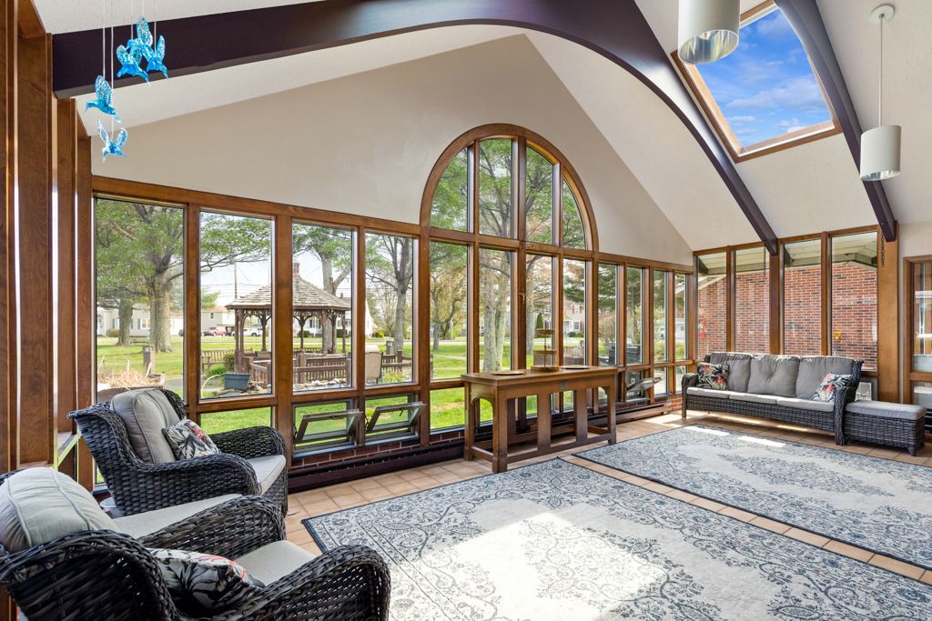 Sunroom with large windows, brown beams, and wicker furniture overlooking a green yard and gazebo.