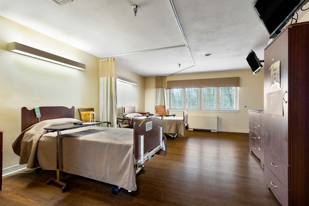 A shared nursing home room with two beds, a cabinet, and a window.