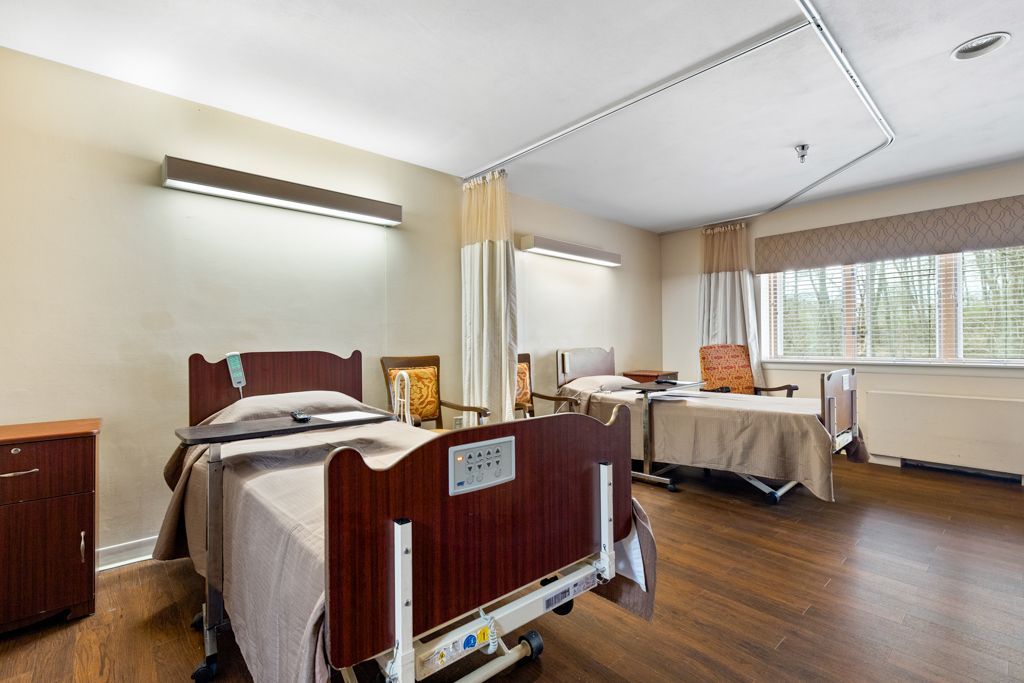 Nursing home room with two beds, wood floors, beige walls, and a window with blinds.