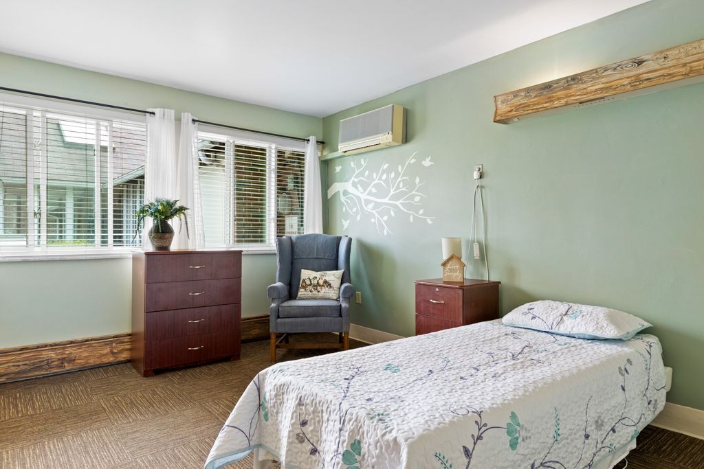 A serene bedroom with a bed, armchair, dresser, and window. Pale green walls and floral decor.