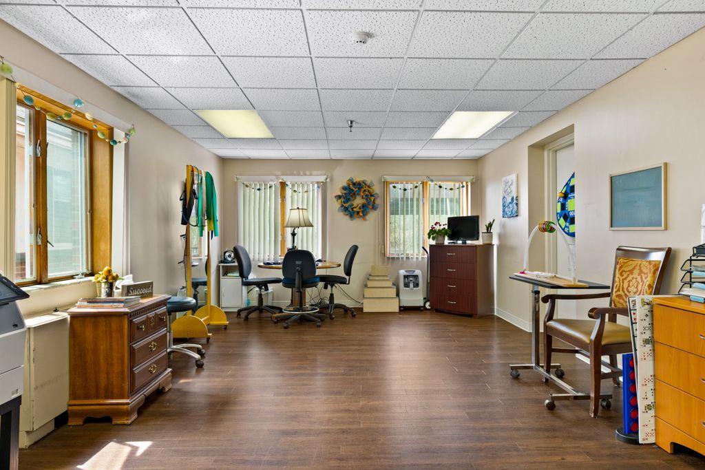 Spacious room with dark wood floor, windows, tables, chairs, and cabinets. Ceiling lights and a television are visible.