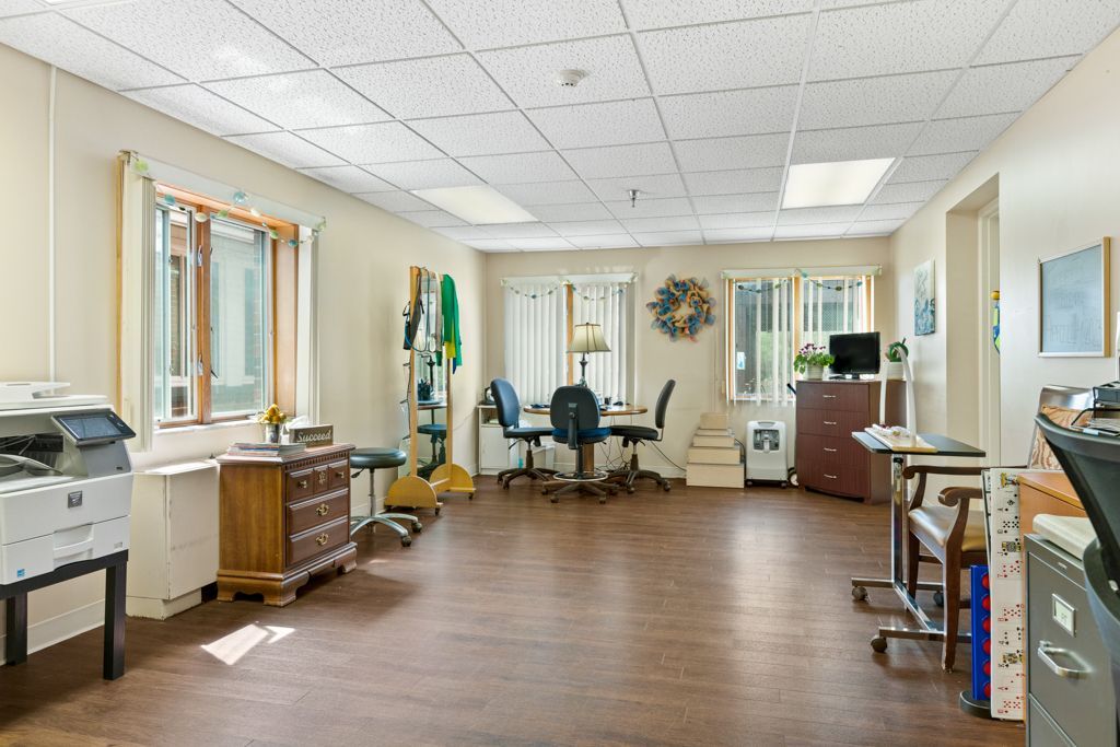 Spacious room with windows, desk, chairs, printer, and wood-look flooring. Neutral-toned walls and ceiling.