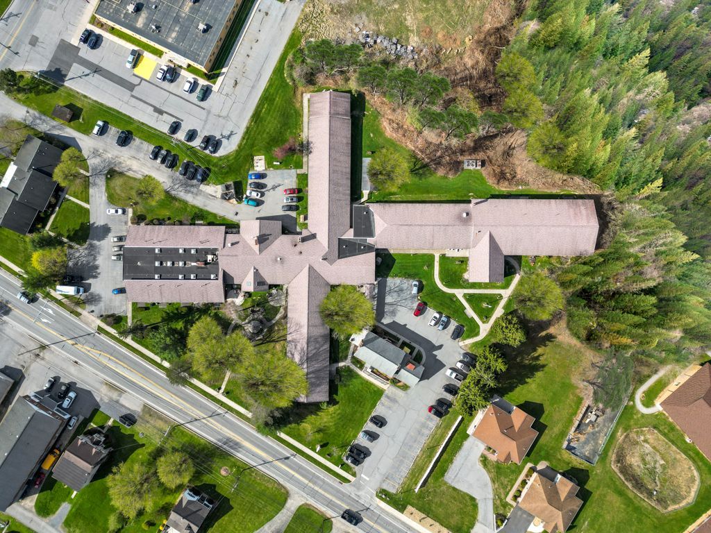 Aerial view of a cross-shaped building with a brown roof surrounded by green grass and trees, and parking lots.